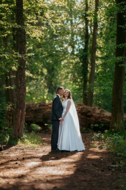 Bir gelin ve damat ormanda öpüşüyor. Gelin beyaz bir elbise giyiyor ve damat bir takım elbise giyiyor. Sahne romantik ve huzurlu, çift doğa ile çevrili.