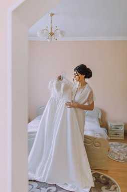 Askıda asılı beyaz bir elbisenin önünde bir gelin duruyor. Elbiseyi tutuyor ve gülümsüyor.