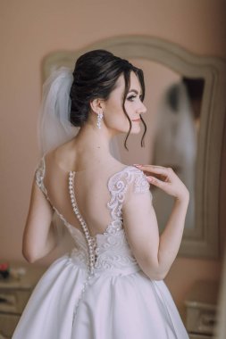 Gelinlikli bir kadın aynanın önünde duruyor. O, dantelli ve beyaz peçeli beyaz bir elbise giyiyor. Elbise uzun ve dalgalı, ve kadının saçı topuz şeklinde.