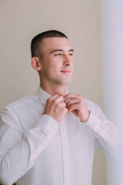 Bir adam resmi bir olaya hazırlanıyor, beyaz bir gömlek giyiyor ve kravatını düzeltiyor.
