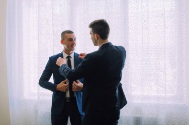 Takım elbiseli iki adam pencerenin önünde dikiliyorlar, biri kravatını düzeltiyor. Erkekler takım elbise ve kravat giydiği için sahne profesyonel ve resmi.