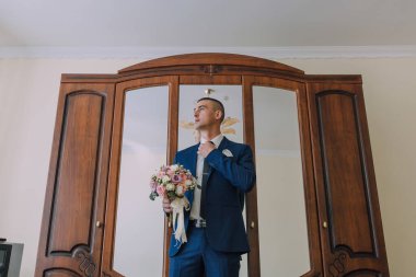 Takım elbiseli bir adam ahşap bir dolabın önünde elinde bir buket çiçekle duruyor.