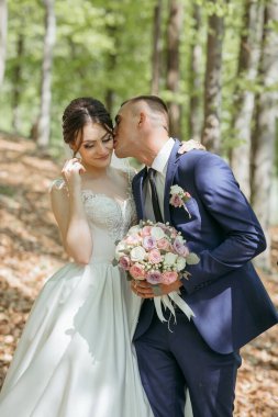 Bir erkek bir buket çiçek tutarken bir kadının yüzünü öper. Sahne, çifti çevreleyen ağaçlarla dolu bir ormanda geçiyor. Sahne romantik ve samimi.