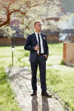 Takım elbiseli ve kravatlı bir adam çimen tarlasında duruyor. Yakasına beyaz bir çiçek takmış.