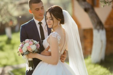 Bir gelin ve damat bahçede birbirini kucaklıyor. Gelin elinde bir buket çiçek tutuyor.