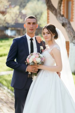 Bir gelin ve damat binanın önünde fotoğraf için poz veriyorlar. Gelin beyaz bir elbise giyiyor ve damat mavi bir takım elbise giyiyor. Gelin elinde bir buket çiçek tutuyor.