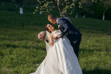 Bir gelin ve damat çimen tarlasında kucaklaşıyor. Gelin beyaz bir elbise giyiyor ve damat mavi bir takım giyiyor.