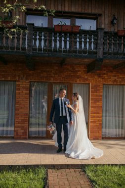 Bir gelin ve damat evin önündeki verandada duruyor. Gelin beyaz bir elbise giyiyor ve damat mavi bir takım elbise giyiyor. El ele tutuşup kameraya gülümsüyorlar.