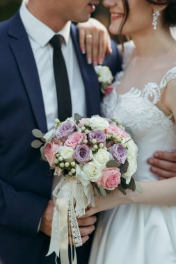 Bir gelin ve damat bir buket çiçek tutarken birbirlerine sarılıyorlar. Gelin beyaz bir elbise giyiyor ve damat mavi bir takım giyiyor.