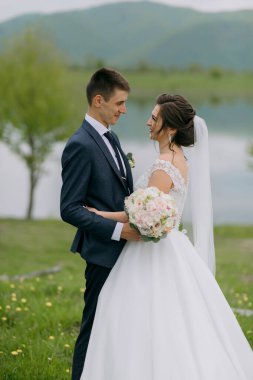Bir gelin ve damat arka planda bir gölle bir tarlada duruyorlar. Gelin bir buket çiçek tutuyor ve damat elini tutuyor.