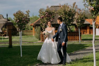 Bir gelin ve damat parkta bir patikada yürüyorlar. Gelin beyaz bir elbise giyiyor ve damat mavi bir takım elbise giyiyor. El ele tutuşuyorlar ve mutlu ve aşık görünüyorlar.