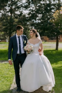 Bir gelin ve damat parkta birlikte yürüyorlar. Gelin beyaz bir elbise giyiyor ve damat mavi bir takım elbise giyiyor. El ele tutuşup kameraya gülümsüyorlar.