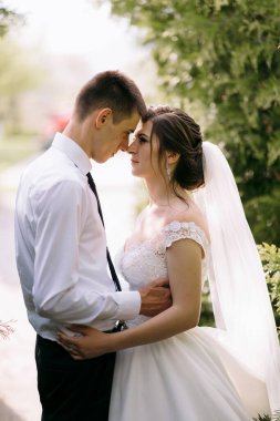 Bir gelin ve damat bahçede birbirini kucaklıyor. Gelin beyaz bir elbise giyiyor ve damat beyaz bir gömlek ve kravat takıyor.