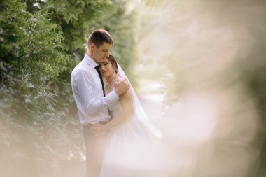 Bir gelin ve damat ormanda sarılıyor. Gelin beyaz bir elbise giyiyor ve damat beyaz bir gömlek giyiyor.