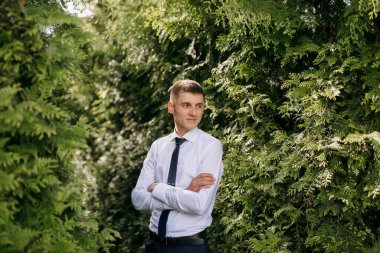 Beyaz gömlekli ve siyah kravatlı bir adam ormanda duruyor. Kameraya ciddi bir ifadeyle bakıyor.