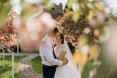 Bir gelin ve damat pembe bir ağacın önünde kucaklaşıyor. Gelin beyaz bir elbise giyiyor ve damat bir takım elbise giyiyor. Sahne romantik ve samimi, aşkı yakalıyor.