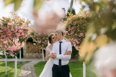 Bir gelin ve damat pembe bir ağacın önünde kucaklaşıyor. Gelin beyaz bir elbise giyiyor ve damat mavi bir kravat takıyor. Sahne romantik ve samimi, aşkı yakalıyor.