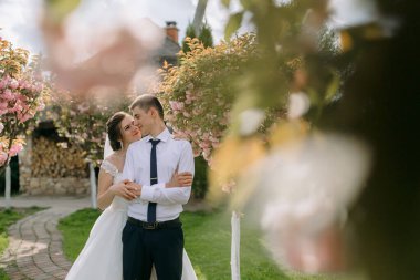 Bir gelin ve damat pembe çiçeklerle bir ağacın önünde duruyorlar. Gelin beyaz bir elbise giyiyor ve damat mavi bir kravat takıyor. Birbirlerini kucaklıyorlar.