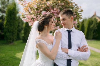 Gelin ve damat parkta, gelin beyaz bir elbise ve damat mavi kravat takıyor. İkisi de gülümsüyor ve birbirlerine bakıyorlar. Muhtemelen düğün günlerinin tadını çıkarıyorlar.