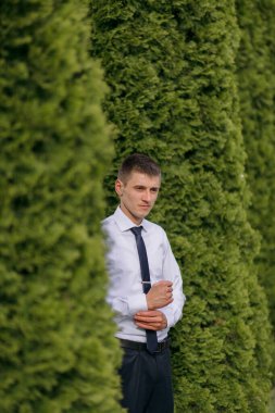 Takım elbiseli ve kravatlı bir adam yeşil bir çitin önünde duruyor. O siyah bir kravat ve beyaz bir gömlek giyiyor.