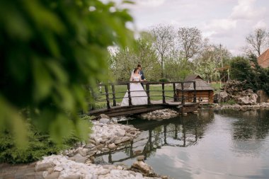 Bir gelin ve damat bir göletin üzerindeki köprüde duruyorlar. Gelin beyaz bir elbise giyiyor ve damat bir takım elbise giyiyor. Sahne huzurlu ve romantik.