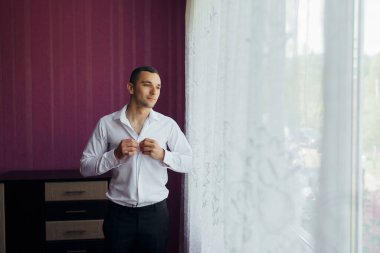 Beyaz gömlekli bir adam pencereden dışarı bakarken yakasını düzeltiyor. Odanın mor bir duvarı ve beyaz bir perdesi var.