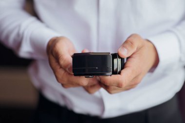 Bir adam elinde bir kamera tutuyor. Kamera siyahtır ve kare şeklindedir. Adam beyaz gömlek ve siyah pantolon giyiyor. Profesyonellik ve odaklanma kavramı