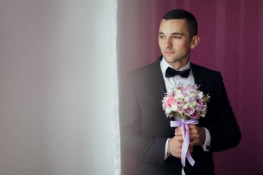Elinde bir buket çiçek tutan takım elbiseli bir adam. Adam siyah bir takım elbise ve siyah bir papyon takıyor. Buket mor ve pembe.