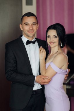 Bir adam ve bir kadın resim için poz veriyorlar. Kadın mor bir elbise giyerken erkek siyah bir takım elbise ve papyon takıyor. İkisinin de yüzünde gülümseme var.