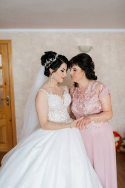 Bir gelin ve annesi bir odada birlikte duruyorlar. Gelin beyaz bir elbise giyiyor ve anne pembe bir elbise giyiyor. Sahne sıcak ve sevgi dolu.