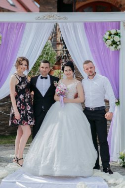 Bir gelin ve damat düğün kemerinin önünde fotoğraf çektirmek için poz veriyorlar. Gelin beyaz bir elbise giyiyor ve damat bir takım elbise giyiyor. Fotoğrafta iki kişi daha var.