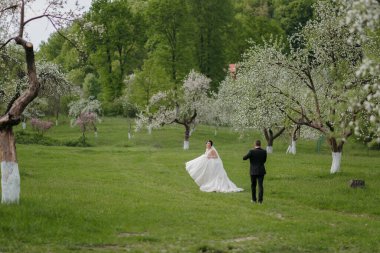 Bir gelin ve damat ağaçların arasında duruyor. Gelin beyaz bir elbise giyiyor ve damat siyah bir takım elbise giyiyor. Huzurlu ve huzurlu bir sahne.