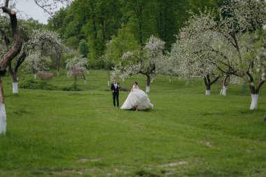 Bir gelin ve damat ağaçların arasında yürüyor. Gelin uzun bir elbise giyiyor ve damat bir takım elbise giyiyor. Sahne huzurlu ve romantik.