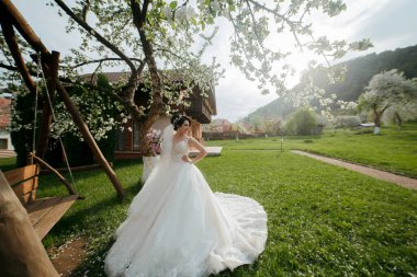 Bir gelin yemyeşil bir tarlada duruyor, beyaz bir elbise giyiyor ve elinde bir buket tutuyor.