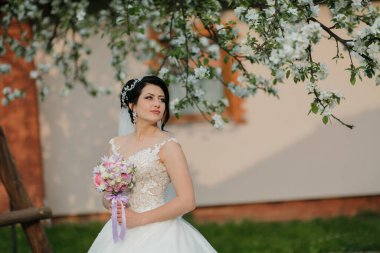 Bir gelin elinde bir buket çiçekle bir binanın önünde duruyor. Beyaz bir elbise ve duvak takıyor.