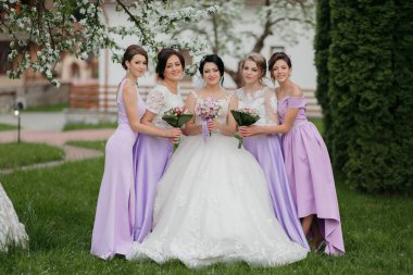 Bir grup kadın parkta fotoğraf çektirmek için poz veriyor. Kadınlardan biri beyaz bir elbise giyiyor ve elinde bir buket çiçek tutuyor. Sahnede mutlu bir sahne var.