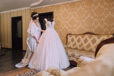 Bir gelin düğün için hazırlanıyor ve bir mankenin üzerinde asılı bir elbisenin önünde duruyor.