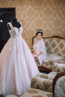 Bir kadın uzun beyaz bir elbisenin önünde duruyor. Elbise bir mankenin üzerinde ve koltuğu ve koltuğu olan bir odada sergileniyor. Odanın sıcak ve davetkar bir atmosferi var.