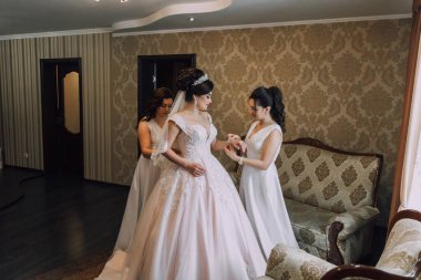 Üç kadın düğün için hazırlanıyor. Biri beyaz bir elbise giyiyor ve diğer ikisi de giyinmesine yardım ediyor.