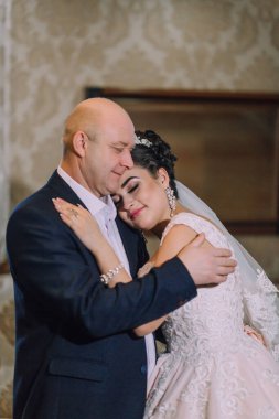 Bir erkek ve bir kadın birbirlerine sarılıyorlar. Adam takım elbise giyiyor ve kadın da gelinlik giyiyor. Sahne sıcak ve sevgi dolu.