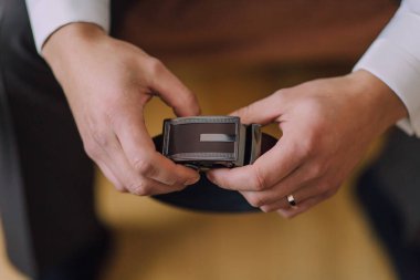 Bir adam elinde kemer tutuyor. Kemer siyah ve altın bir tokası var. Adam beyaz gömlek ve gri pantolon giyiyor.