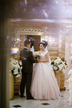 Gelin ve damat aynanın önünde duruyorlar, gelin elinde buket tutuyor. Sahne mutlu ve kutlama dolu, çift düğünlerine hazırlanıyor.