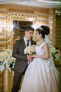 Gelin ve damat aynanın önünde duruyorlar, gelin elinde buket tutuyor. Sahne mutlu ve romantik.