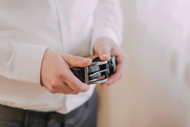 Bir adam elinde bir kemer tutuyor. Kemer siyah ve gümüş bir tokası var. Adam beyaz gömlek ve gri pantolon giyiyor. Formalite ve profesyonellik kavramı