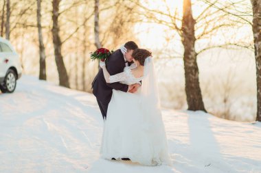 Bir gelin ve damat, arka planda bir arabayla karda kucaklaşıyorlar. Sahne sıcak ve romantiktir. Çift güzel bir kış ortamında düğünlerini kutluyor.
