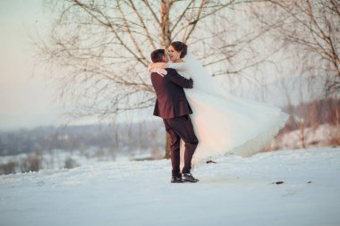 Gelin ve damat karda kucaklaşıyorlar, gelin bir buket tutuyor. Sahne karlı bir manzarada geçiyor, arka planda bir ağaç var. Sahne neşeli ve romantik.