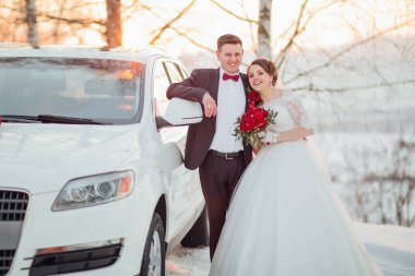 Bir gelin ve damat beyaz bir arabanın önünde poz veriyorlar. Gelin beyaz bir elbise giyiyor ve damat bir takım elbise giyiyor. Araba karda park halinde.