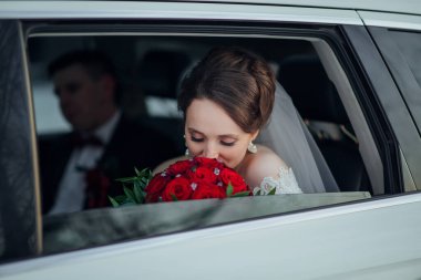Bir gelin, elinde bir buket kırmızı gülle arabanın arkasında oturuyor. Pencereden dışarı bakıyor ve dalgın bir ruh hali içinde.