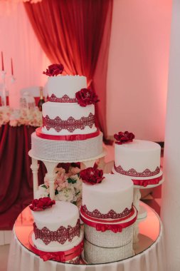 Kırmızı çiçekler ve beyaz kremalı çok katlı bir düğün pastası. Pasta gümüş bir tepsiye konur ve kırmızı perdelerle çevrilidir.