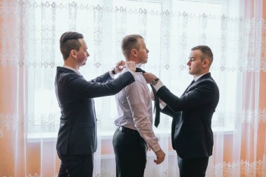 Üç adam takım elbise ve kravat takıyor. Biri diğerinin kravatını düzeltiyor.